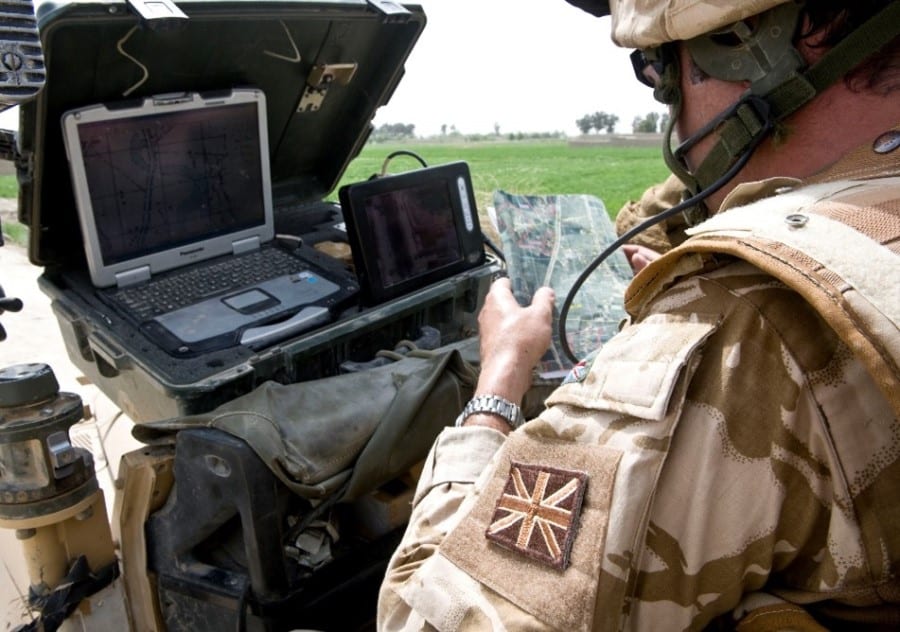 Military personnel wearing brown camo in the field working on a field computer and talking on comms.