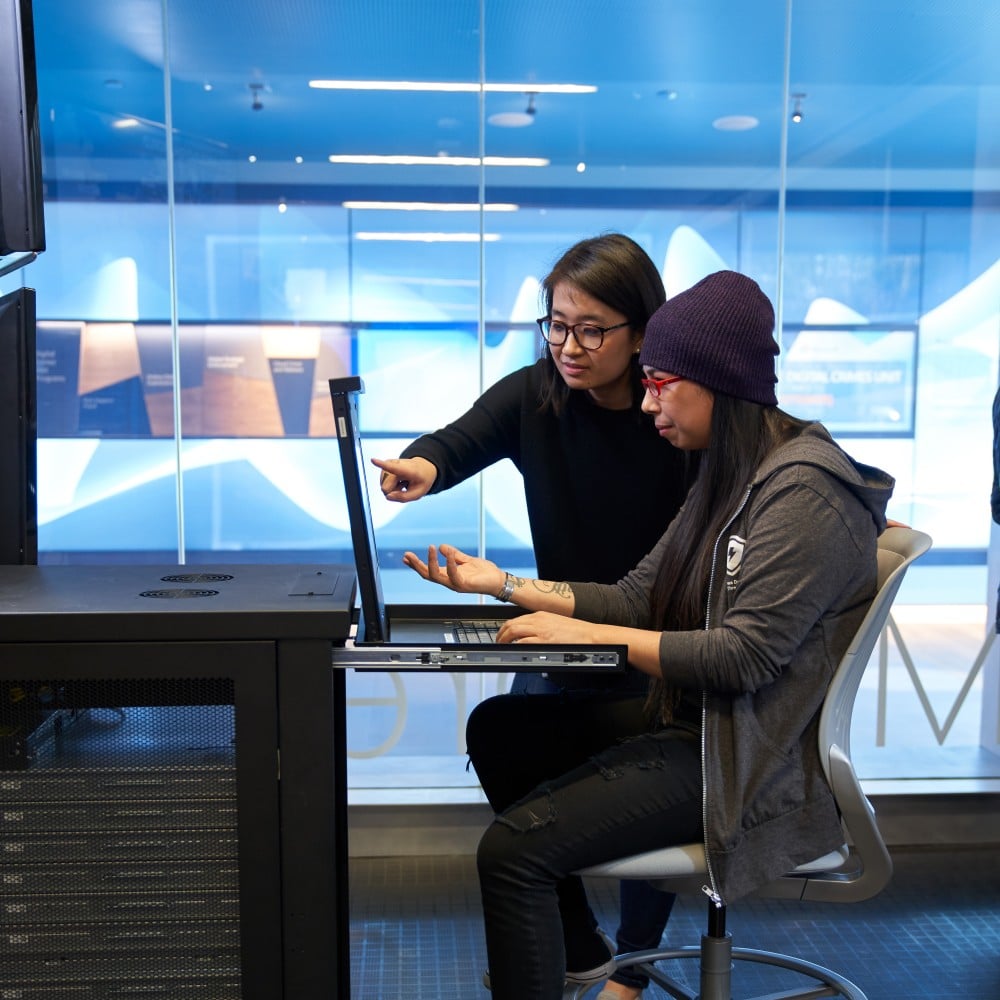 Two women working on a network server station inside a secure room, discussing and pointing to something on the screen.