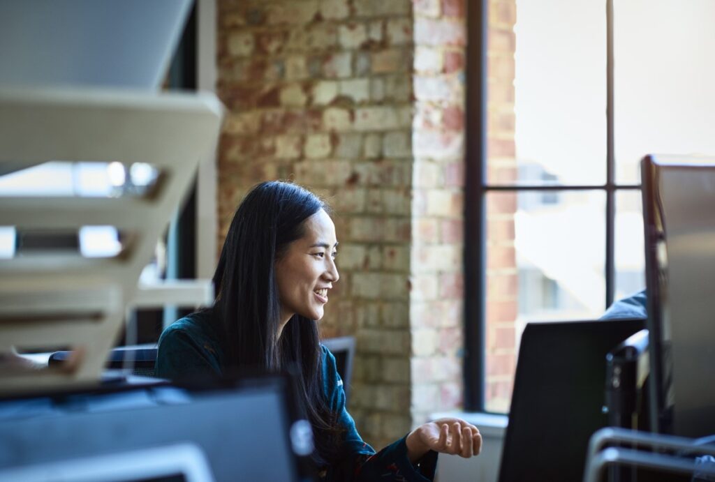 Office worker, smiling and looking away, in contemporary studio with bare brick walls, sitting in front of a desktop computer.