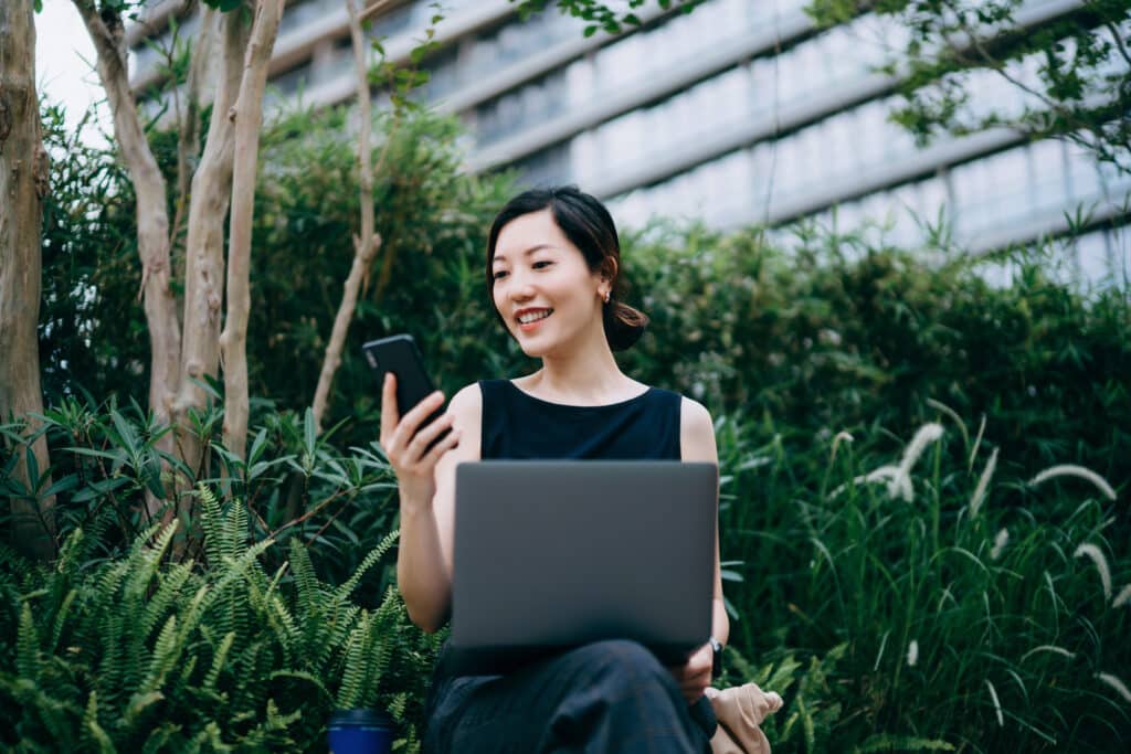 Woman working with laptop outdoors in urban office park, surrounded by green plants.